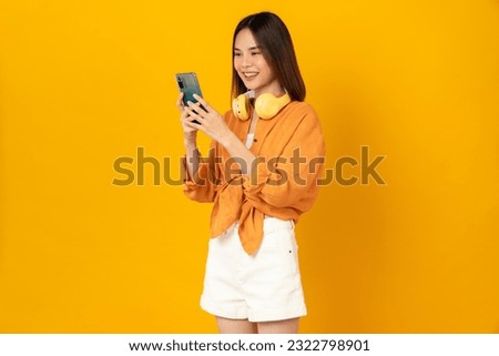 Cheerful young Asian woman in headphones listening to music and enjoy favourite playlist application on smartphone with dancing on orange background.