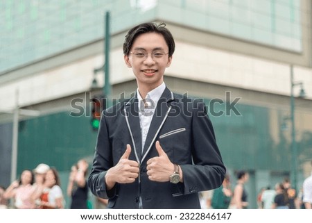 A young chinese man in his early 20s makes a composed double thumbs up sign while walking outside the city plaza.