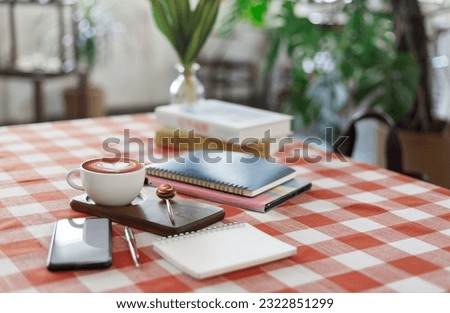 Coffee cup with note book and smart phone, reading book put on table with red and white tablecloth, people relax and take a break with coffee in cafe during a day before start job again