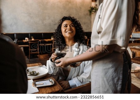 Young smiling african woman using smartphone while paying restaurant bill with contactless payment during dinner party with friends Royalty-Free Stock Photo #2322856651