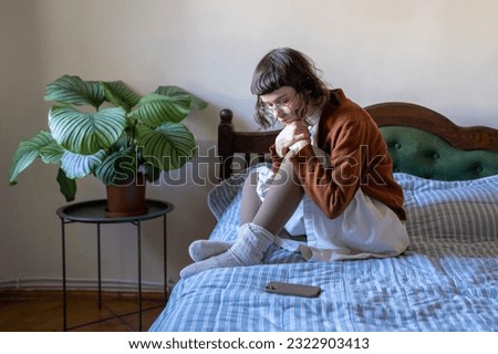 Sad depressed teenage girl in eyeglasses sitting on bed waits for call from boyfriend in difficult relations. No answer phone message. Loneliness without cellphone sms. Network internet addiction