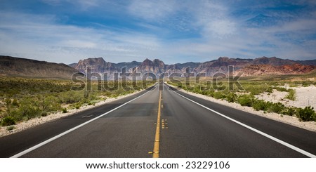 Straight paved road running towards the Red Rock Canyon National Reserve in Nevada, USA with bushes on the sides, hills in the background.