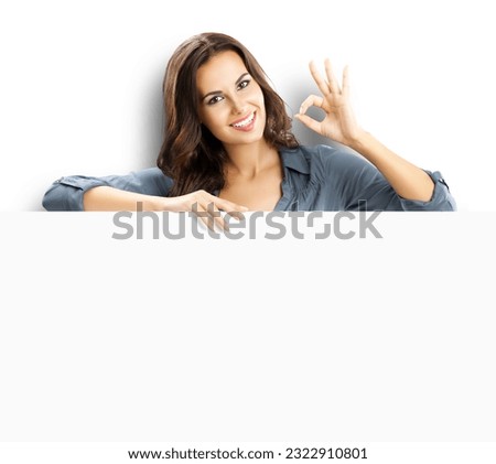 Happy smiling brunette young woman showing okay ok gesture, standing behind, peeping from blank banner or mock up signboard with copy space, isolated over white background.