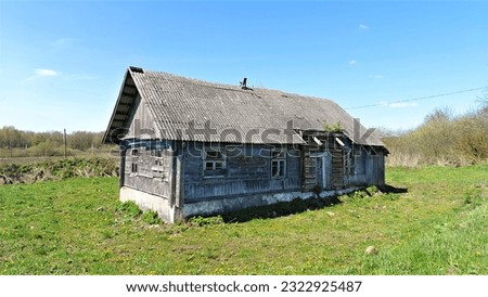 On a grassy lawn stands an old wooden house with boarded-up windows and a slate roof. Next to it are trees and shrubs. Behind the house is a farm field and a road, and behind it is a forest. Sunny Royalty-Free Stock Photo #2322925487