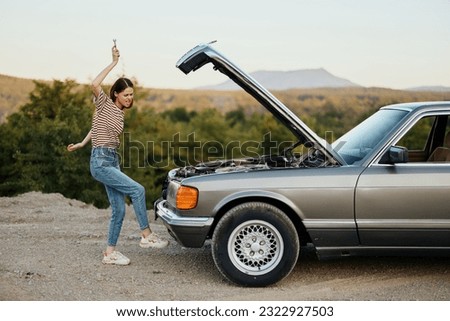 A woman opens the hood of a broken down car and tries to find the cause of the breakdown on a road trip alone and kicks the car in anger Royalty-Free Stock Photo #2322927503