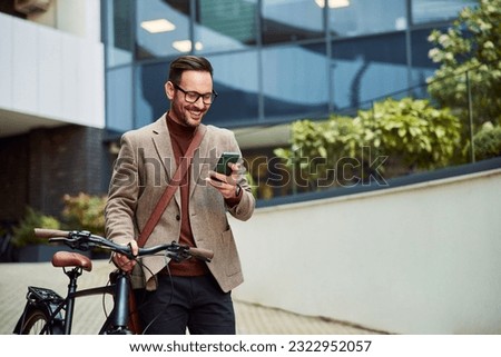 A cheerful businessman with glasses on reading text messages on a smart phone and pushing a bicycle.