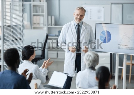 Happy experienced clinician in lab coat standing in front of audience clapping their hands while congratulating him on successful report Royalty-Free Stock Photo #2322953871