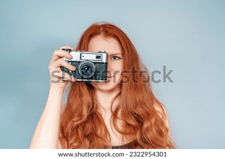 Studio photo. Red-haired woman with camera in black dress. Gray background.