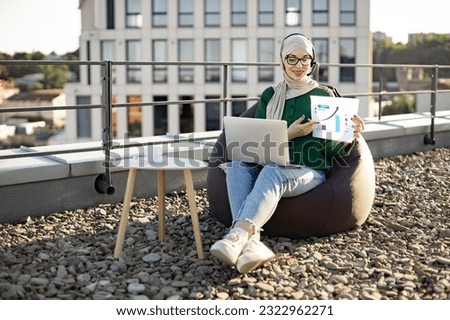 Young arabian female in casual clothes and hijab giving online presentation using headset and computer outside office. Remote manager brainstorming about statistics shown by infographics on terrace.