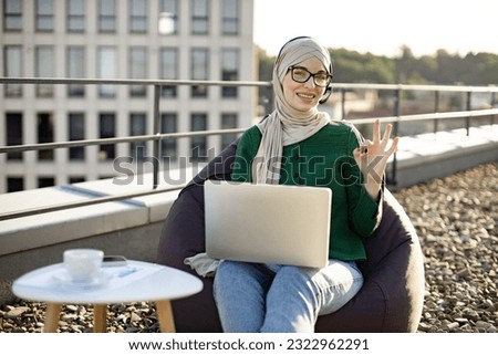 Beautiful arabian lady in hijab posing with portable computer and headset in cozy beanbag on roof terrace. Well-organised young employee enjoying open air while staying available during midday sign ok