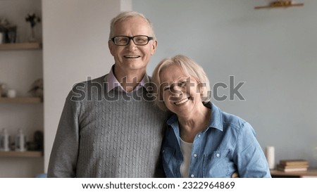 Portrait of smiling senior couple in love hugging pose in living room look at camera. Medical insurance cover for seniors ad, happy lifelong marriage, harmonic and warm relationships between spouses