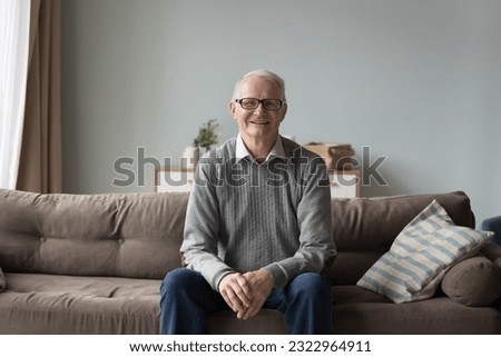 Calm retired elderly man in glasses and casual clothes resting alone seated on comfortable couch in living room smile looking at camera at own or rented home. Baby-boomer generation person portrait Royalty-Free Stock Photo #2322964911