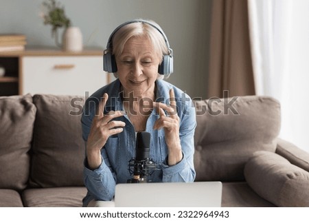 Elderly female blogger wear headphones speaking into professional microphone lead webinar or streaming using laptop, records podcast. Senior radio host, speaker holding program on-air, broadcasting Royalty-Free Stock Photo #2322964943