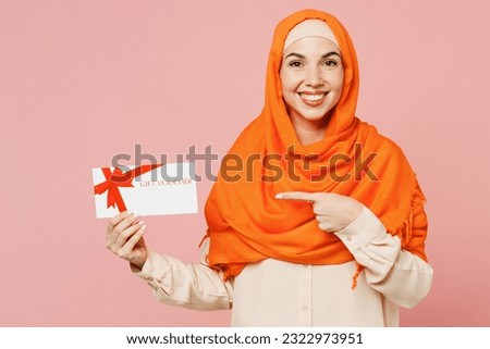 Young arabian asian muslim woman wear orange abaya hijab hold point on store gift certificate coupon voucher card isolated on plain pink background studio. Uae middle eastern islam religious concept