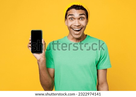 Young surprised man of African American ethnicity wears casual clothes green t-shirt hat hold in hand use mobile cell phone with blank screen workspace area isolated on plain yellow background studio