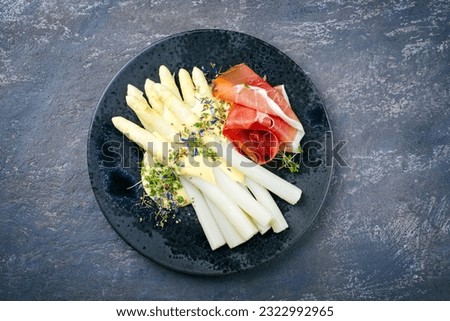 Modern style traditional steamed white asparagus with cured ham and garnished with sauce hollandaise served as top view on a Nordic design plate with copy space  Royalty-Free Stock Photo #2322992965