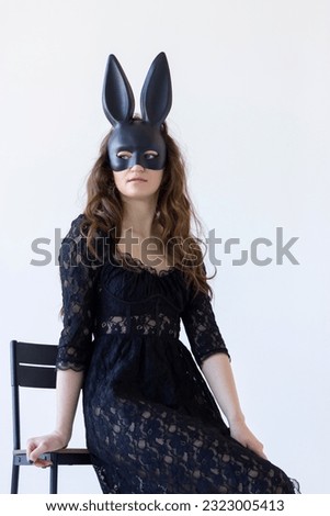 Young Woman Wearing Rabbit, Hare Black Mask On Face Sitting on chair white background, wall. Vertical Plane. High quality photo