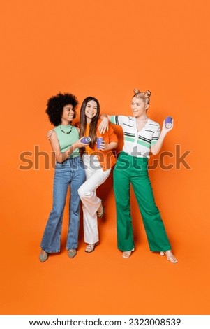 Full length of positive and multiethnic teenage girlfriends in stylish outfits holding canner drink and posing on orange background, trendy outfits and fashion-forward looks Royalty-Free Stock Photo #2323008539