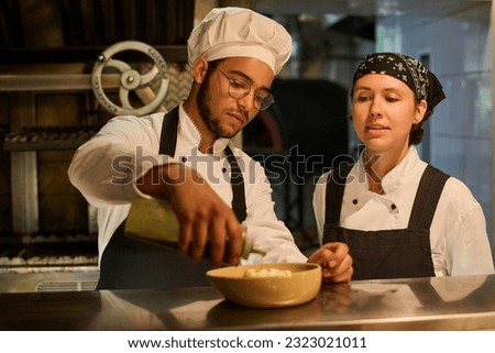 Young serious male chef in uniform sprinkling sauce on top of fresh salad