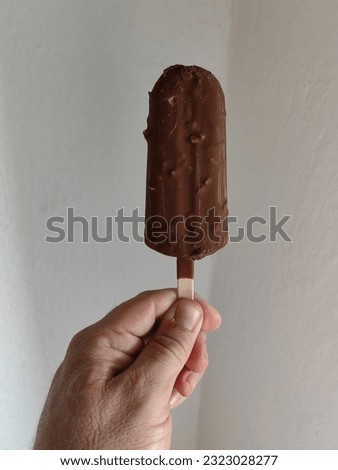 Man' s hand holding dark brown ice cream, against white wall background. Summertime  season.