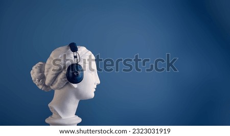The profile of the plaster head of a graceful Aphrodite in huge blue audio headphones, against a blue background. Copy space view. The concept of listening to music, audiobooks, podcasts, lectures.
