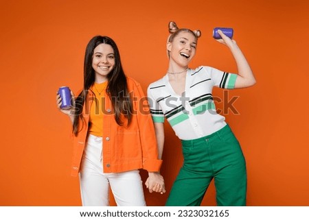 Cheerful blonde and brunette teenage girlfriends in casual outfits holding drink in tin cans and holding hands while standing on orange background, fashionable girls with sense of style Royalty-Free Stock Photo #2323032165