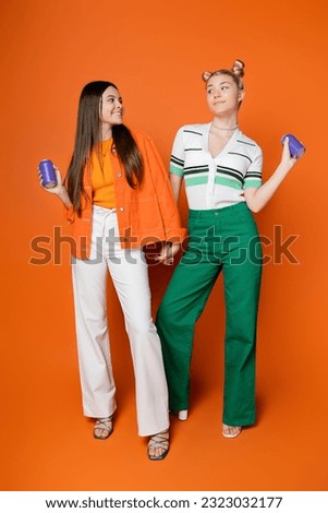 Full length of smiling and stylish brunette teenager holding hand of blonde girlfriend and drink in tin can and standing together on orange background, fashionable girls with sense of style Royalty-Free Stock Photo #2323032177