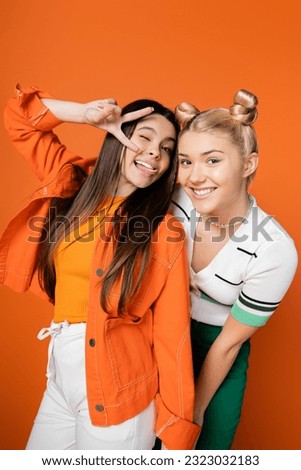 Cheerful blonde and brunette teenage girlfriends with bright makeup and stylish outfits gesturing and posing at camera while standing isolated on orange, fashionable girls with sense of style Royalty-Free Stock Photo #2323032183