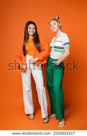 Full length of cheerful blonde and brunette teenage models in trendy outfits posing and looking at camera on orange background, fashionable girls with sense of style, friendship and bonding