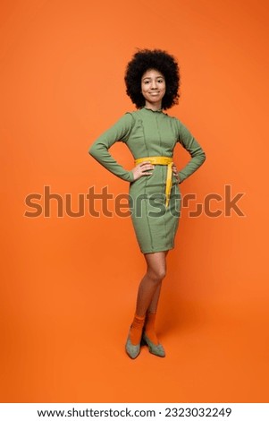 Full length of smiling and fashionable african american teen girl with bold makeup posing in green dress while standing on orange background, teenage fashion and generation z concept