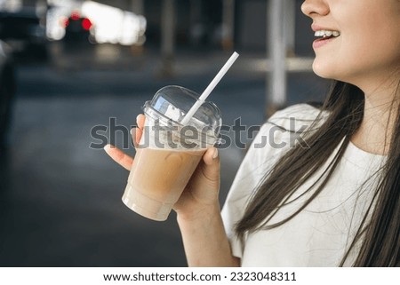 Close-up, a disposable cup of iced coffee in female hands on a blurred background of a parking lot, cooling a summer drink. Royalty-Free Stock Photo #2323048311