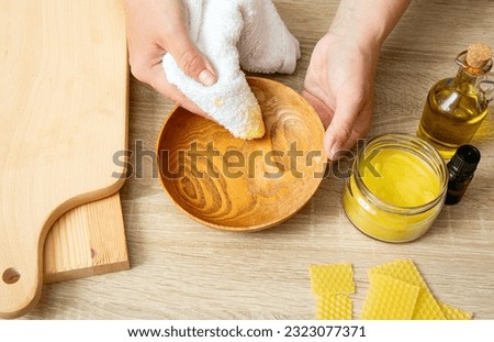 Woman hands apply homemade beeswax wood treatment polish to restore natural wood bowl color. Beeswax, olive oil and essential oil, soft cloth and mixture in glass jar. Polishing wood.  Royalty-Free Stock Photo #2323077371