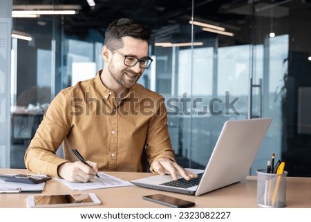 Smiling male businessman sits in the office at the table, works with documents and talks with conducts a business meeting online. Royalty-Free Stock Photo #2323082227