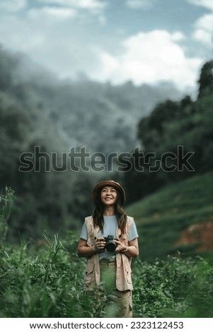Smiling tourist asian woman photographer holding camera in meadow with mountains and fog in the background, journey for sightseeing in nature, Travel and hobby concept