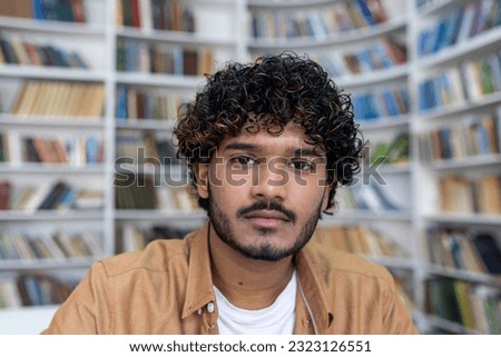 Close-up photo. Portrait of a young Indian male student sitting in the library near the bookshelves and looking seriously into the camera.
