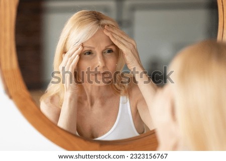 Senior woman looking at mirror and touching forehead, upset aged lady examining fine lines on her face, suffering skin aging, selective focus on reflection Royalty-Free Stock Photo #2323156767