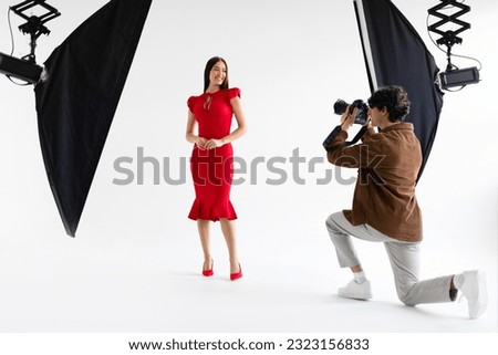 Fashion photoshoot. Professional photographer taking photos of beautiful female model in elegant red dress, posing on white background in photo studio with modern lighting equipment