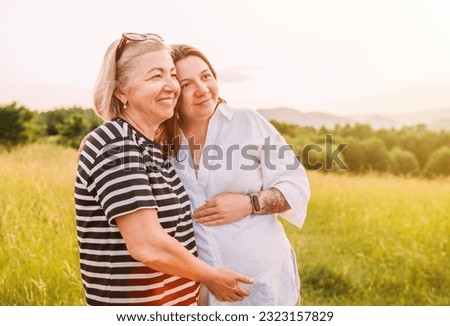 Portrait of a sincerely smiling young pregnant woman dressed in light summer clothes embracing with mother in evening sunset hours. Woman's health, human in the nature concept image. Royalty-Free Stock Photo #2323157829