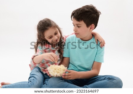 Lovely little girl hugging her older brother while watching movie or cartoons, sitting together isolated on white studio background. Kids. Entertainment. Leisure. Lifestyle. Happy family relationships