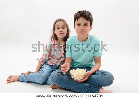 Adorable cheerful happy kids, pre teen boy and little girl eating popcorn, watching movie or cartoons, looking toward the camera, sitting together on isolated white studio background. Family leisures