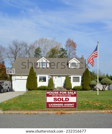American flag pole Real Estate sold (another success let us help you buy sell your next home) sign suburban cape cod style home autumn fall day residential neighborhood blue sky clouds USA