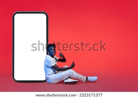Online assistant and chatbot concept. Positive millennial african american guy in white casual outfit sitting by huge phone with blank screen and showing thumb up, mockup, copy space