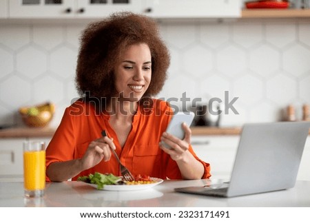 Smiling black woman using smartphone and laptop while having lunch in kitchen at home, cheerful african american female sitting at table, browsing new app on mobile phone and eating food, free space