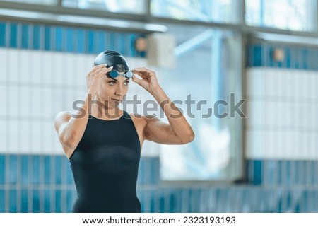 Portrait of a woman, a professional swimmer in black one-piece swimwear and a black cap, looking at the camera and putting on swimming goggles. Royalty-Free Stock Photo #2323193193