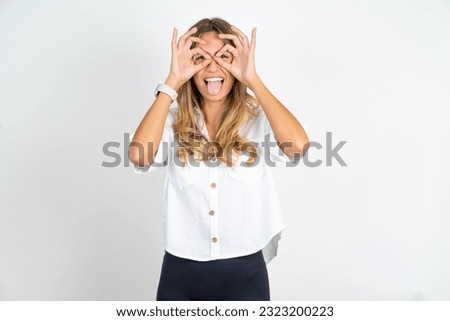 Young caucasian business woman wearing white shirt over white background doing ok gesture like binoculars sticking tongue out, eyes looking through fingers. Crazy expression.