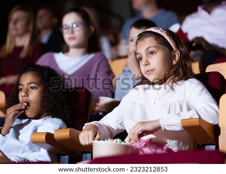Interested preteen girl enjoying watching movie and eating popcorn with her african american friend in picture theater