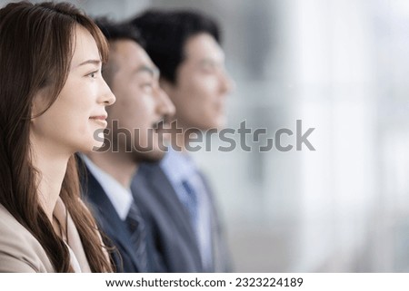 Three Japanese men and women in suits lined up in a large office, smiling and staring into the distance. Perfect for a job offer or career change image. Copy space available. Royalty-Free Stock Photo #2323224189