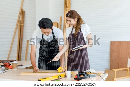 Asian professional male and female lover couple carpenter worker in apron using measuring tape measure wooden sticks on workbench with equipment in home decoration building workshop Royalty-Free Stock Photo #2323240369
