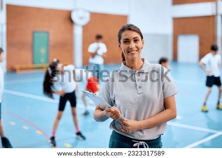 Happy physical education teacher during exercise class at school gymnasium looking at camera. Her students are in the background.