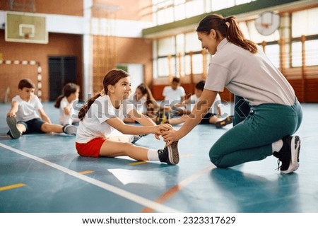 Happy schoolgirl stretching her leg with teacher's assistance on physical education class at school gym. Royalty-Free Stock Photo #2323317629
